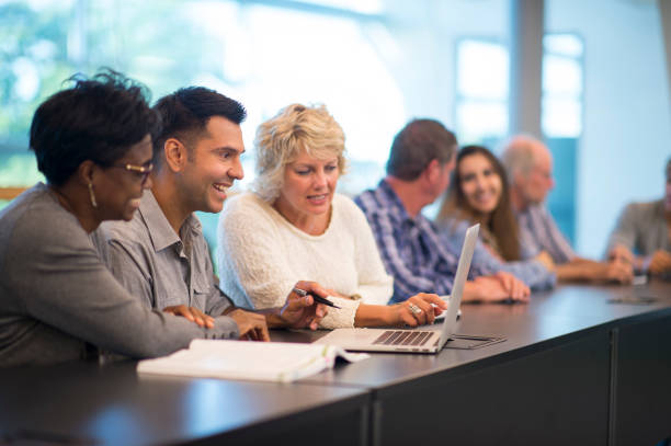 Several members of a university faculty are sitting together in a boardroom. Three of them are looking at one colleague's laptop.